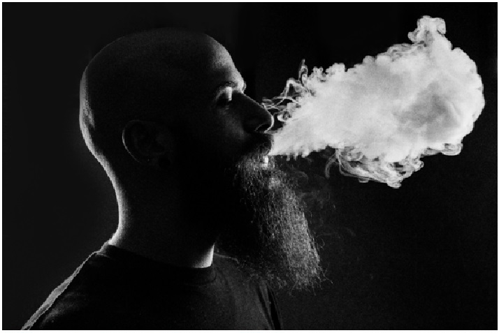 Man with a beard exhaling a vape cloud in black and white.