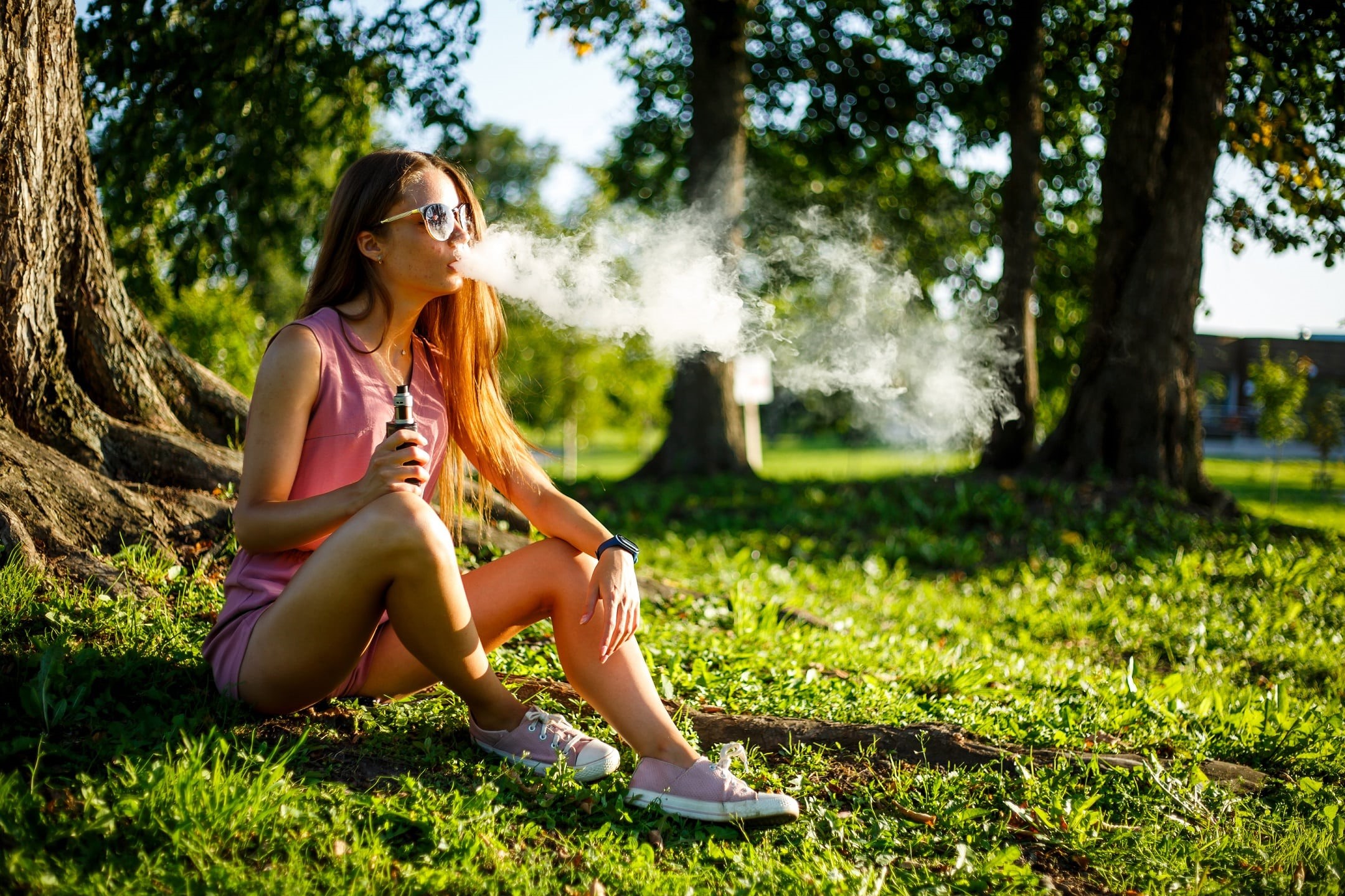 Young pretty sporty caucasian girl in a pink jumpsuit and sunglasses smoking an electronic cigarette or vape in the park on a sunny day in summer